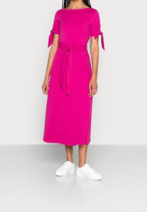 Vrouw in een felroze midi-jurk met korte mouwen en strikken, een taille riem en witte sneakers, staande op een grijze vloer.