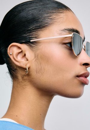Profil einer Frau mit zurückgekämmt geglättetem Haar, die goldene Creolen- und Steckohrringe sowie goldgerahmte, reflektierende Sonnenbrillen trägt.