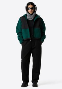 Giacca in pile verde con dettagli neri, cappuccio grigio, maglietta nera e pantaloni cargo neri. Gli accessori includono occhiali da sole scuri e scarpe nere.