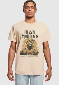 Beige bomulds T-shirt med et grafisk design af en kædret figur og tekst, der siger "Iron Maiden World Tour 1984-85".