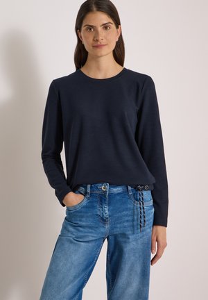 Femme aux longs cheveux foncés, portant une chemise bleu marine à manches longues et un jean bleu, une main dans la poche, sur un fond uni.