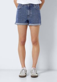 Pantalones cortos de mezclilla en azul claro, con un diseño clásico de cinco bolsillos, dobladillos vuelta y un acabado desgastado. Combinados con zapatillas gris y blancas.