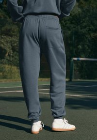 Grijze joggingbroek met elastische boorden en tailleband, met een achterkant zak. Gecombineerd met witte sneakers op een groene tennisbaan.