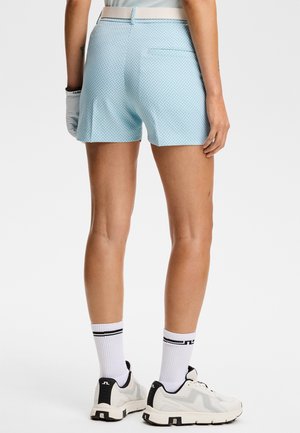 Femme portant un short léger bleu à motifs, une ceinture blanche, un gant de golf blanc, des chaussettes blanches et des baskets blanches, debout, dos tourné.