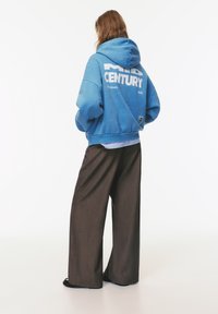 Felpa con cappuccio blu con testo bianco "MID CENTURY" sul retro, abbinata a pantaloni larghi marroni. Tessuto morbido con una vestibilità rilassata e un design oversize.