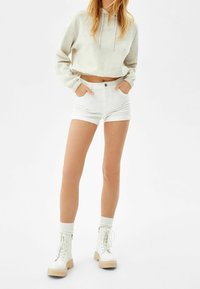 Mujer vistiendo un cropped de sudadera gris claro, shorts blancos de talle alto, calcetines blancos y botas de tobillo blancas con cordones, de pie contra un fondo blanco.