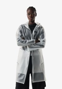 Woman with braided hair wears a translucent long raincoat with front pockets and hood over black outfit, standing with arms crossed.