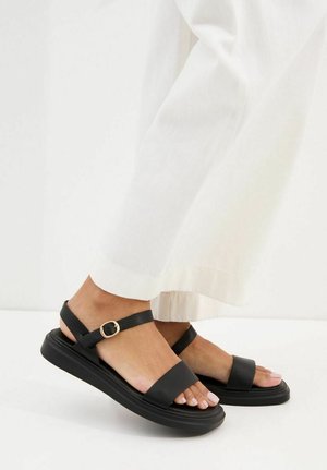 Stopy w czarnych skórzanych sandałach z paskami wokół kostki i złotymi klamrami, w połączeniu z luźnymi białymi spodniami na jednolitym białym tle.