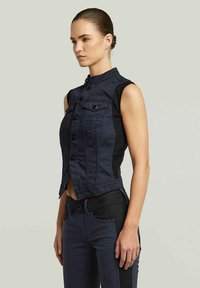 Gilet in denim con collo alto, pannelli laterali neri e chiusure a bottoni. Colore blu scuro con due tasche sul petto e design corto.