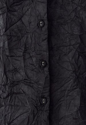 Chemise noire à boutons, fabriquée en tissu froissé, avec quatre boutons noirs et une surface texturée présentant des plis et des rides visibles.