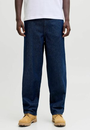 Person trägt locker sitzende dunkelblaue Jeans, hellbraune Schnürstiefel und ein weißes, geripptes Shirt vor einem schlichten, hellen Hintergrund.