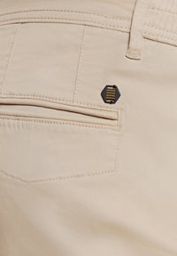 Beige broek van een gestructureerde stof, met een achterzak en een klein zwart en goud hexagonaal logo detail. Netjes zichtbare stiksels.