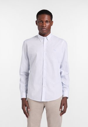 Giovane uomo che indossa una camicia azzurra a maniche lunghe con bottoni e pantaloni beige, in piedi contro uno sfondo bianco.