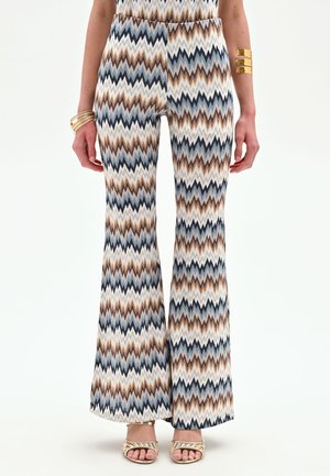 Donna che indossa pantaloni a gamba larga con motivo a zigzag in blu, marrone, nero e bianco, abbinati a bracciali dorati e tacchi aperti dorati.