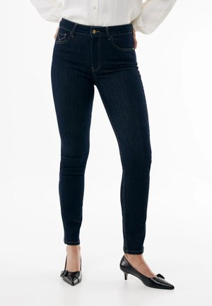 Kvinde iført mørkeblå skinny jeans og sorte lave hælede spidse sko, stående med hænderne på hoften mod en hvid baggrund.