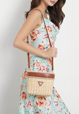 Nainen kukallisessa hihattomassa mekossa kantaa beige- ja kermaista, nahkaosilla varustettua olkalaukkua.