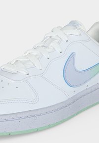 Λευκό αθλητικό παπούτσι Nike με ιριδίζον swoosh, λευκά κορδόνια και ανοιχτοπράσινη, με κηλίδες σόλα, από πλάγια όψη σε λευκό φόντο.