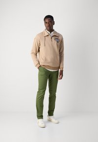Beige hoodie with half-zip and pocket, featuring colorful embroidery; paired with slim-fit green pants and white sneakers with accents.