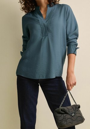 Kvinde iført en teal langærmet bluse og mørke jeans, der holder en grå quiltet håndtaske med en sølvkædehank i den ene hånd.