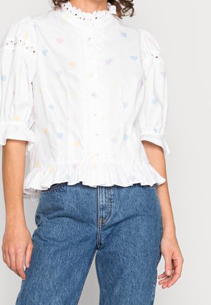 Blouse blanche avec motifs de cœurs pastel, boutonnage festonné à l’avant, bas et manches à volants, portée avec un jean bleu taille haute.