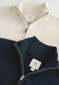 Două pulovere cu fermoar, din tricot elastic pliat, unul bej și unul bleumarin, fiecare având o etichetă „NEXT” în interiorul gulerului.