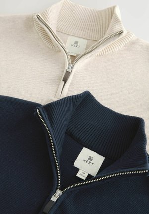 Două pulovere cu fermoar, din tricot elastic pliat, unul bej și unul bleumarin, fiecare având o etichetă „NEXT” în interiorul gulerului.