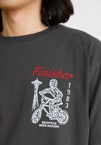 T-shirt gris foncé avec un graphique blanc d'un cycliste BMX, la skyline de Seattle et un texte rouge "Finisher 1983" indiquant un style vintage.