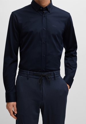 Mand iført en tætsiddende mørk navy skjorte med knapper på kraven og matchende teksturerede navy bukser med snøre i taljen.