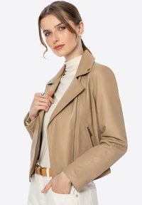 Beige Lederjacke mit Reverskragen, Reißverschlusstaschen und taillierter Passform. Tragen über einem cremfarbenen Rollkragenpullover und weißen Hosen.