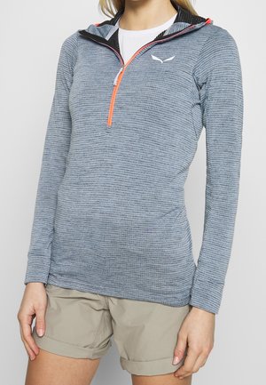 Femme portant un pull texturé gris clair à manches longues avec une fermeture éclair orange et un short beige retroussé sur un fond uni.