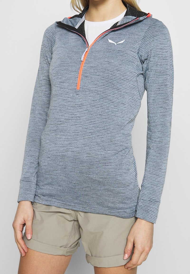 Femme portant un pull texturé gris clair à manches longues avec une fermeture éclair orange et un short beige retroussé sur un fond uni.