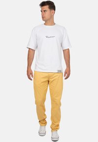 T-shirt bianco a maniche corte con logo nero, abbinato a pantaloni gialli. Le calzature presentano sneakers bianche con dettagli rossi.
