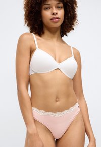 Balts polsterētais krūšturis ar regulējamām siksniņām, apvienots ar gaiši rozā biksītēm ar mežģīņu apdari jostasvietā. Gluda auduma tekstūra.