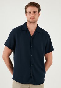 Korte mouwen marineblauw overhemd met knopen in een soepele stof. Kenmerkt zich door een klassieke kraag en een rechte zoom. Subtiele detail van een zijzak aan de voorkant.