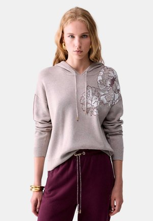 Donna bionda che indossa una felpa con cappuccio beige chiaro con ricamo floreale sulla spalla e pantaloni viola scuro con coulisse, accessoriata con braccialetti dorati.