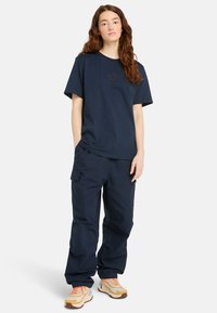 Marineblaues Baumwoll-T-Shirt mit kleinem Logo, kombiniert mit marineblauen Cargo-Hosen mit Seitentaschen. Getragen mit mehrfarbigen Turnschuhen.