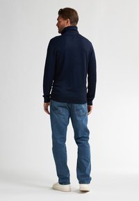 Marineblauwe pullover met een hoge kraag, geribbelde mouwen en zoom, gecombineerd met lichtblauwe jeans en witte sneakers, zichtbaar vanuit de achterkant.