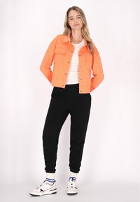 Jachetă scurtă de culoare portocalie intensă, cu două buzunare pe piept, asortată cu pantaloni jogger negri și adidași albi cu accente negre și albastre.