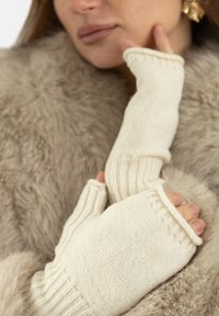 Rękawiczki bezpalcowe w kremowym kolorze, wykonane z dzianiny o prążkowanej fakturze i przeszyciach. Ręce modelki ułożone na miękkim, jasnym futrzanym tle.