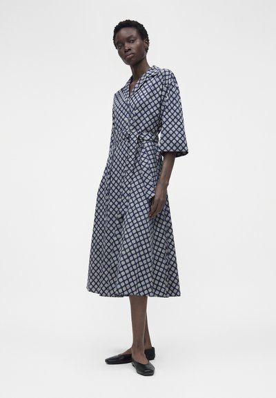 Ψηλή γυναίκα που φοράει ένα μπλε σκούρο φόρεμα μήκους μίντι με σχέδια, μανίκια μέχρι τον αγκώνα και μαύρα μπαλαρίνια, στέκεται μπροστά σε απλό λευκό φόντο.