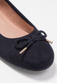 Nahaufnahme einer navyblauen Wildleder-Balletflats mit geflochtenem Bogen und goldenen Spitzen auf einem weißen Hintergrund.