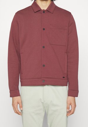 Mand iført en vinrød skjortejakke med brystlomme og sidelommer, kombineret med lyse beige bukser, stående mod en hvid baggrund.