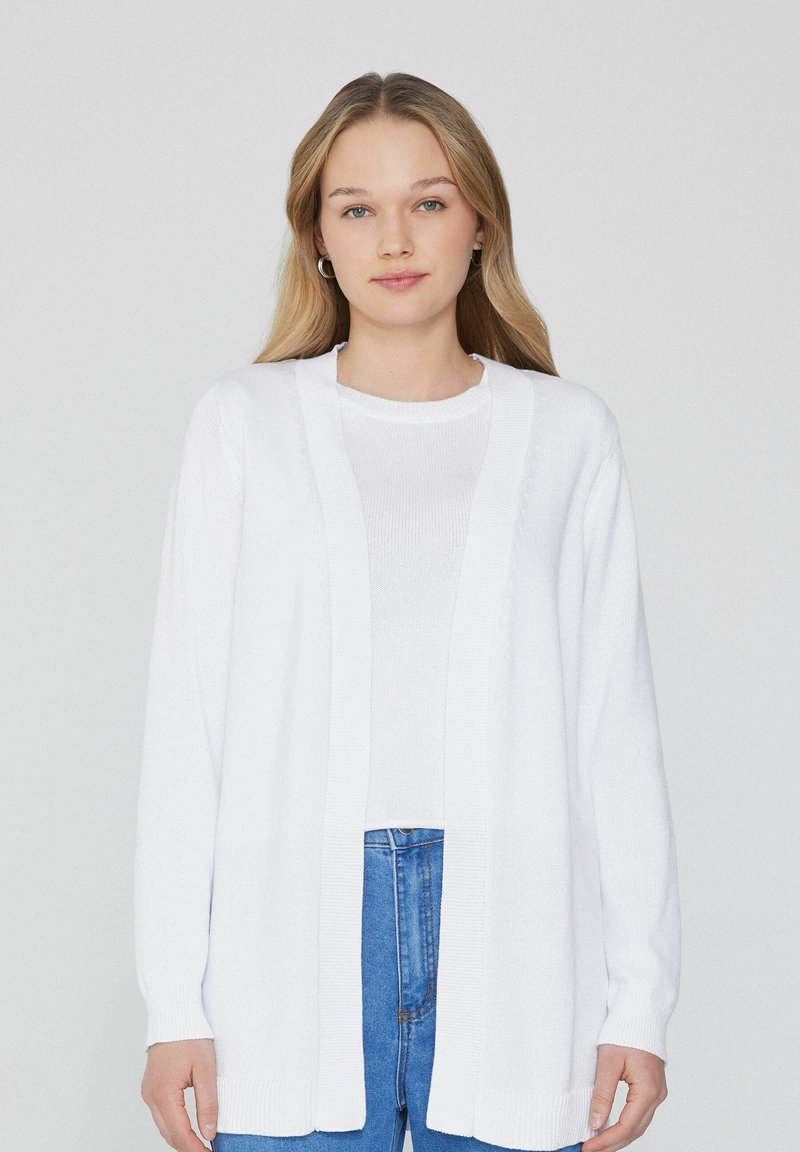 Cardigan blanc avec un design à ouverture frontale, manches longues, poignets et ourlet côtelés, fabriqué en tissu tricot léger. Assorti à un haut blanc.