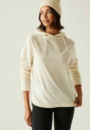 Vrouw met een crèmekleurige hoodie en gebreide muts, gecombineerd met zwarte legging, staand tegen een effen lichte achtergrond.