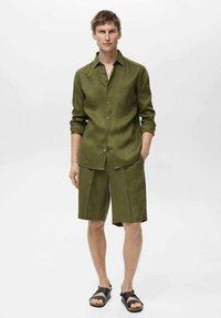 Camicia in lino verde oliva con maniche lunghe e bottoni, abbinata a pantaloni corti fino al ginocchio dello stesso colore. Sandali neri completano l'outfit.