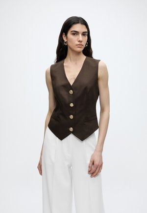 Kvinde iført en ærmeløs mørkebrun vest med guldknapper og hvide bukser med vide ben stående mod en ensfarvet baggrund.