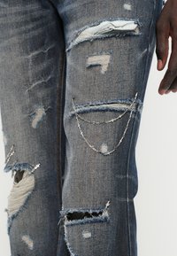 Σκούρα μπλε φθαρμένα τζιν με σκισμένα σημεία, ξέφτια στις άκρες και μια ασημένια αλυσίδα που διακοσμεί την περιοχή γύρω από το γόνατο.