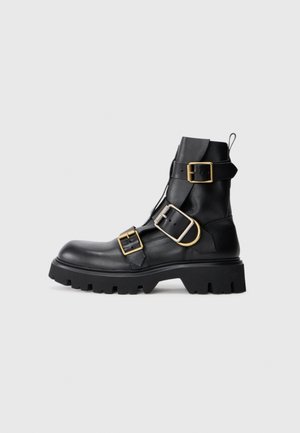 Schwarze Leder-Stiefelette mit mehreren goldenen Schnallen, dickem schwarzen Gummisohle, abgerundetem Zehenbereich und verstärktem Absatz. Mit einer Zuglasche ausgestattet.