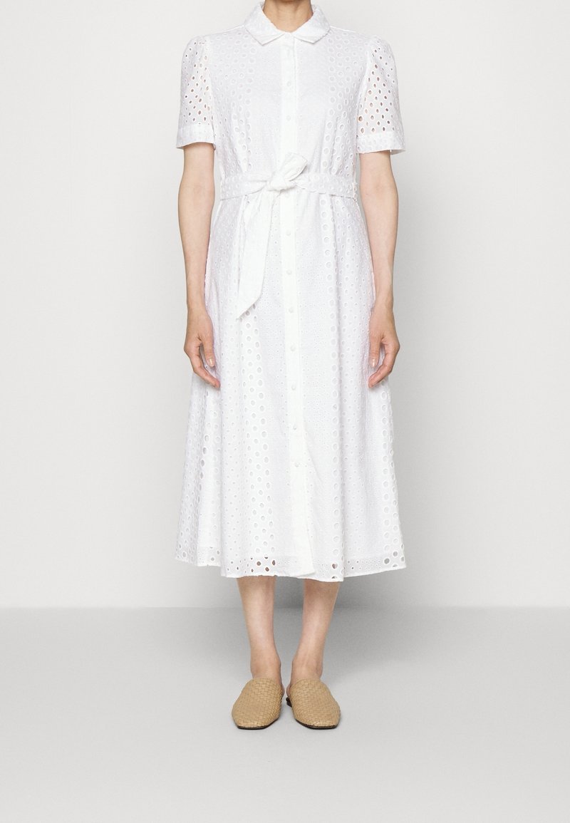Robe midi blanche avec manches courtes bouffantes, tissu ajouré, fermeture à boutons sur le devant, ceinture nouée à la taille, et chaussures beiges tissées à enfiler.
