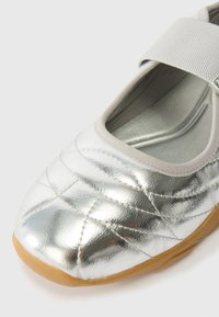 Silver quilted ballet flats with a grey elastic strap, featuring a smooth texture and a flexible rubber sole.